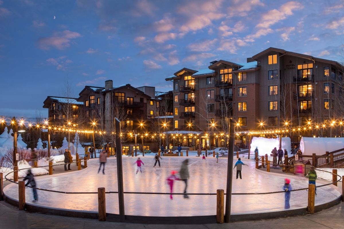 People ice skating on an outdoor rink in front of a multistory lodge at dusk.