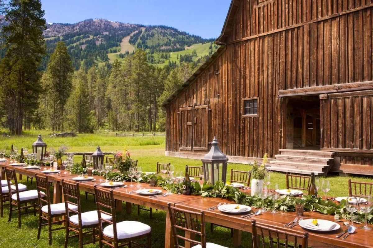 Long banquet table with place settings arranged on a lawn in front of a rustic wooden barn and mountain backdrop.