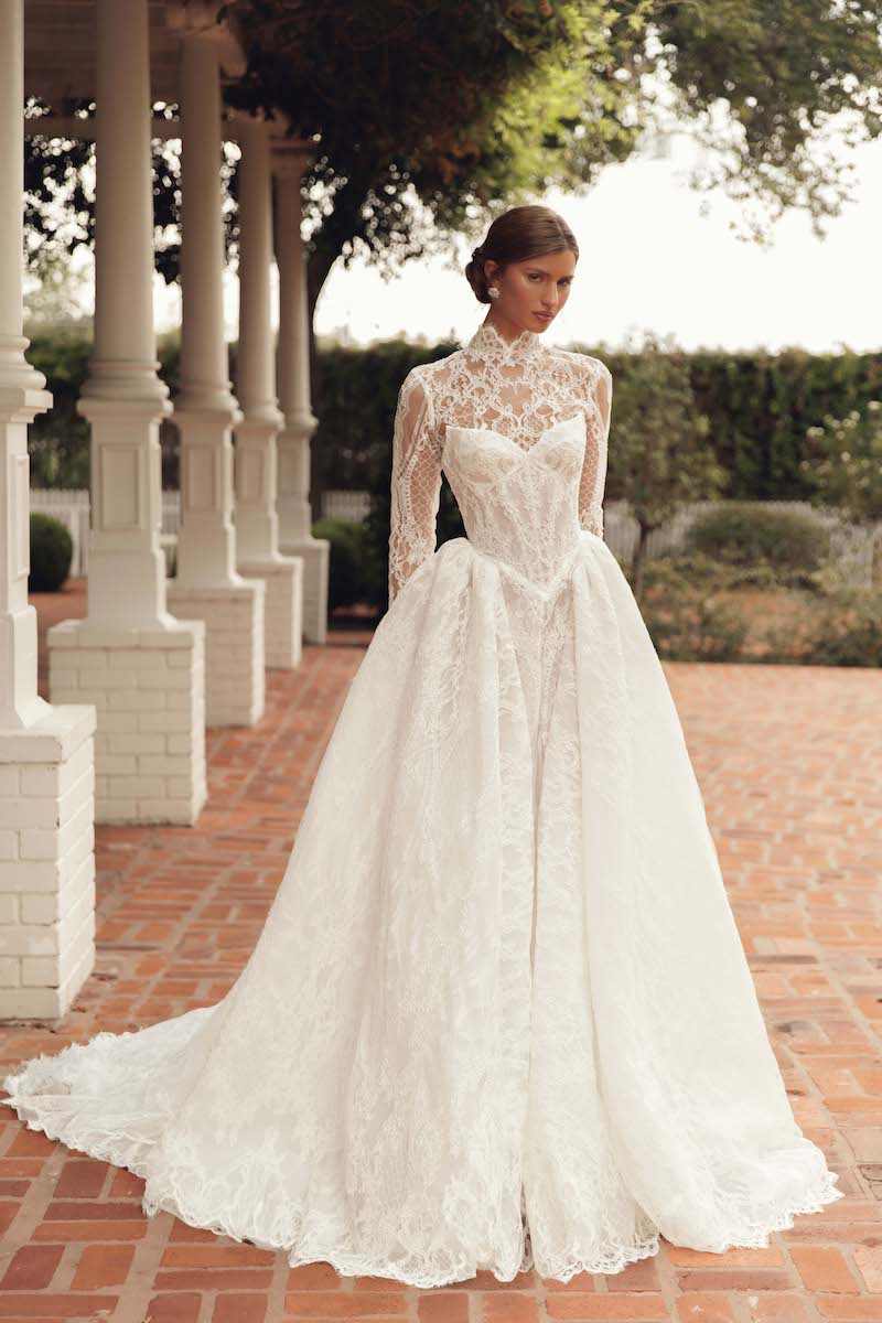 A woman wearing a long-sleeved, high-neck lace wedding dress with a full skirt and train.