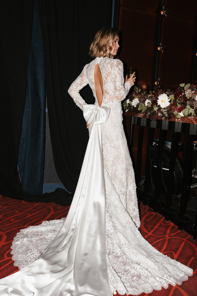 A woman in a long-sleeved, floor-length lace wedding dress with an open back and a large white bow at the waist.