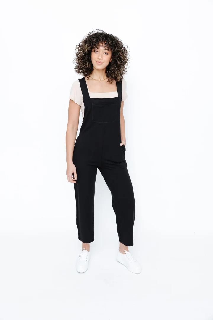 A woman wearing black overalls over a light-colored t-shirt and white sneakers.
