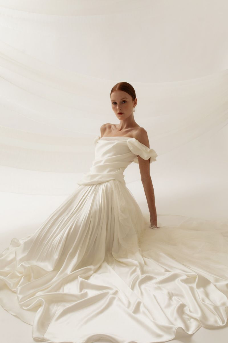 A woman in an off-the-shoulder white gown with a long, draped skirt sitting on the floor.
