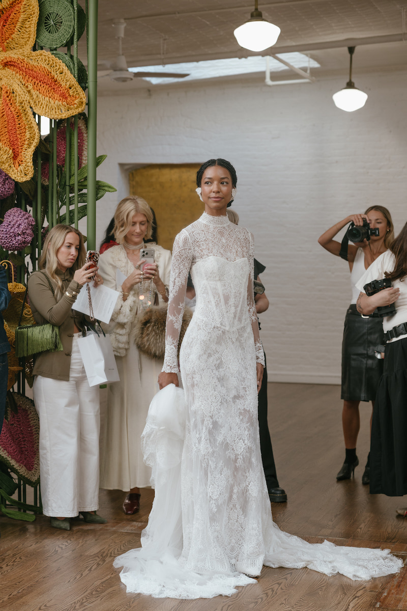 A model walks a runway wearing a long-sleeved, high-neck white lace wedding gown with a train, as guests photograph her.
