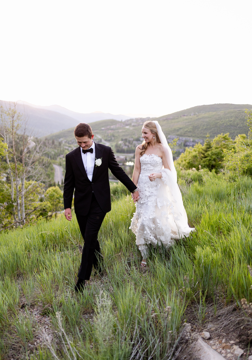 A bride and groom walk through a grassy field with mountains in the background.