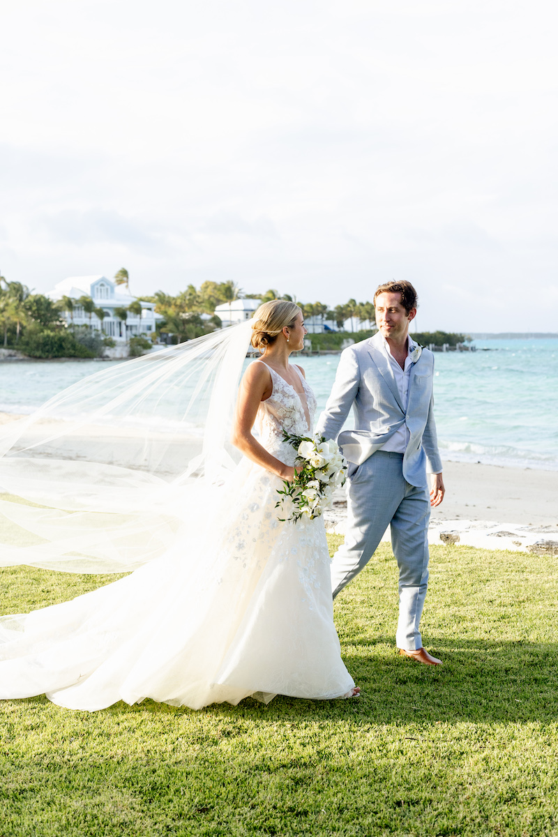 A bride in a white wedding gown and a groom in a light blue suit walk together along the water’s edge.