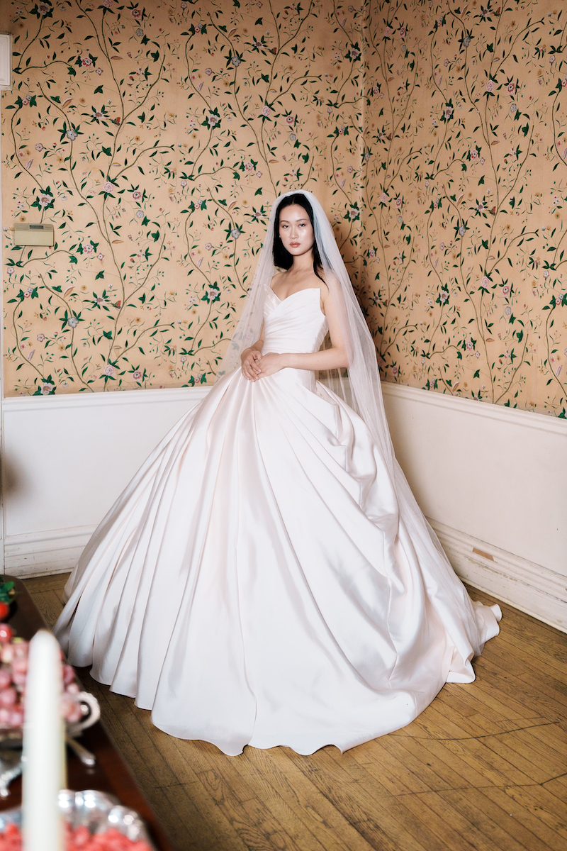 A bride in a strapless ballgown and veil standing in a corner with floral wallpaper.