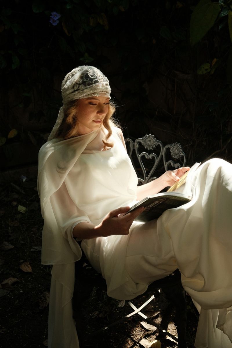 A woman in a white dress and beaded headpiece sitting in a chair and reading a book.