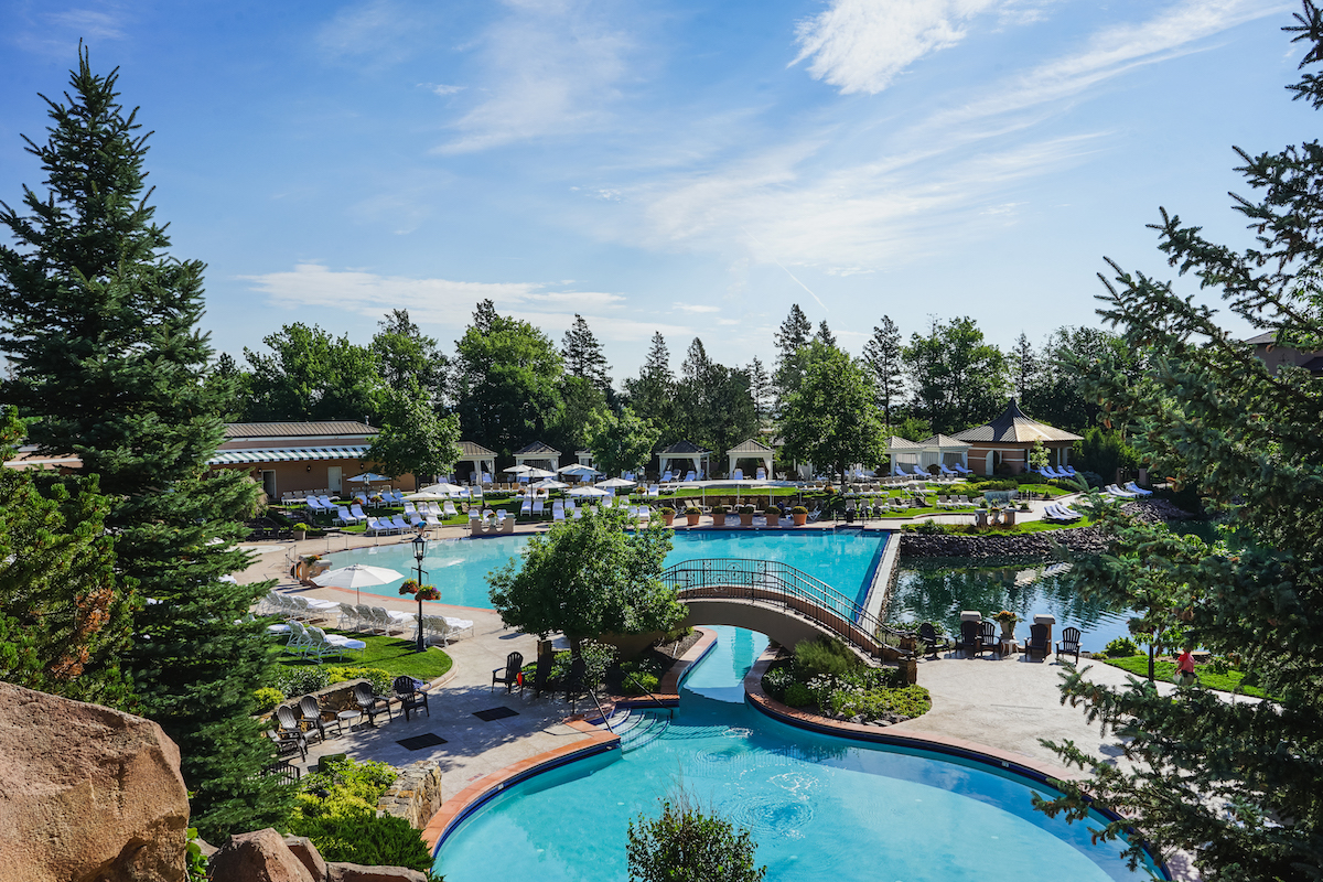 An outdoor swimming pool complex with arched stone bridges connecting turquoise pools, surrounded by trees and lounge chairs.