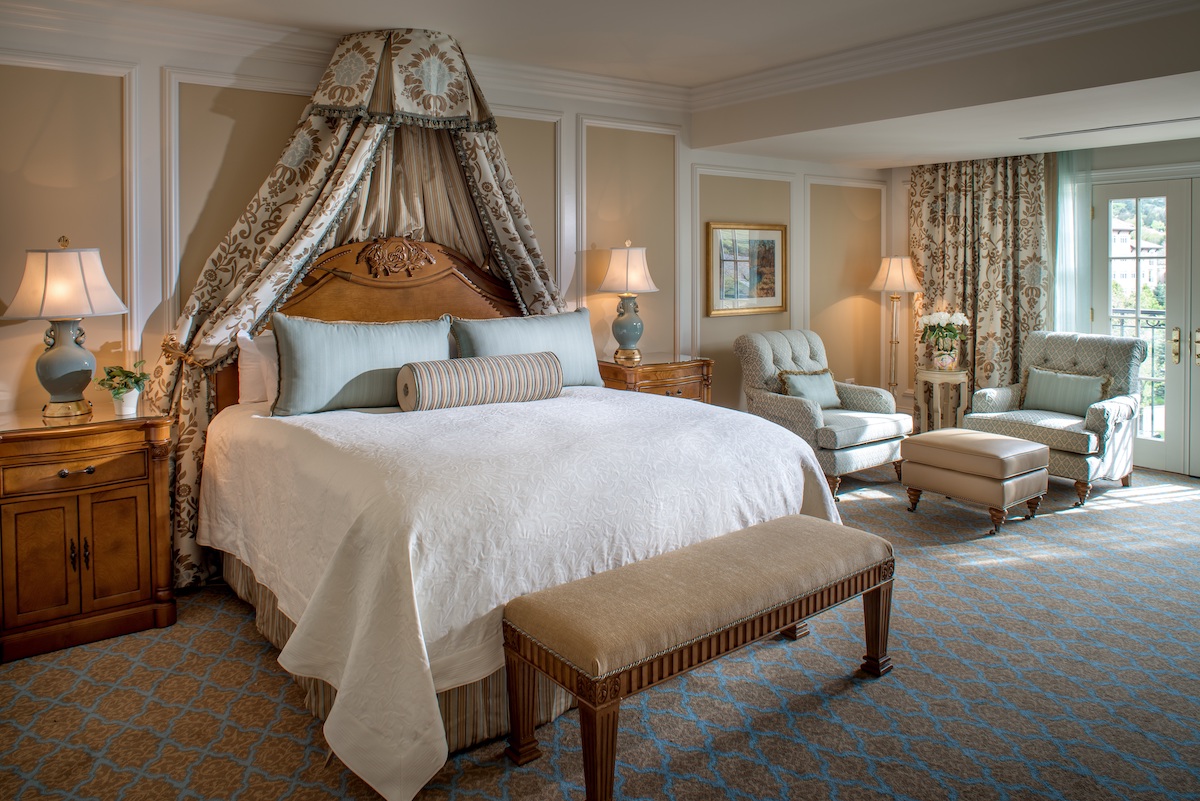 A hotel room with a king bed featuring a decorative canopy, a bench at the foot of the bed, and a sitting area with two armchairs and an ottoman by a window.