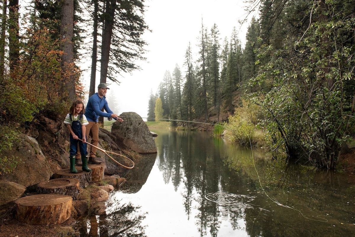 An adult and a child fly fishing together on a riverbank.