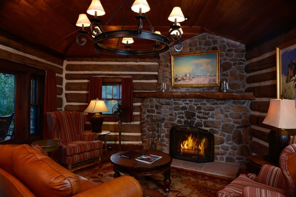 A rustic log cabin living room featuring a stone fireplace, a wrought iron chandelier, and leather seating.
