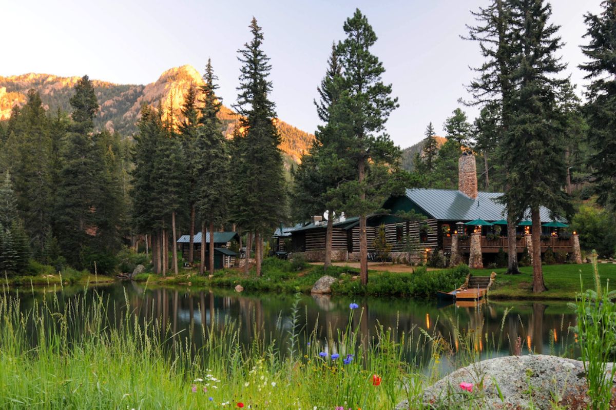 A log cabin nestled among pine trees beside a pond, with a mountain peak in the background.