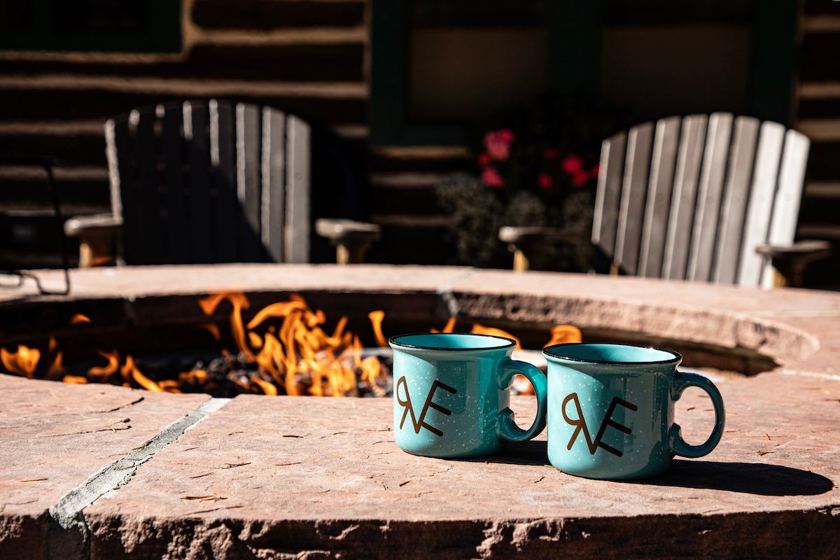 Two light blue mugs on a stone ledge in front of a fire pit, with two wooden chairs in the background.