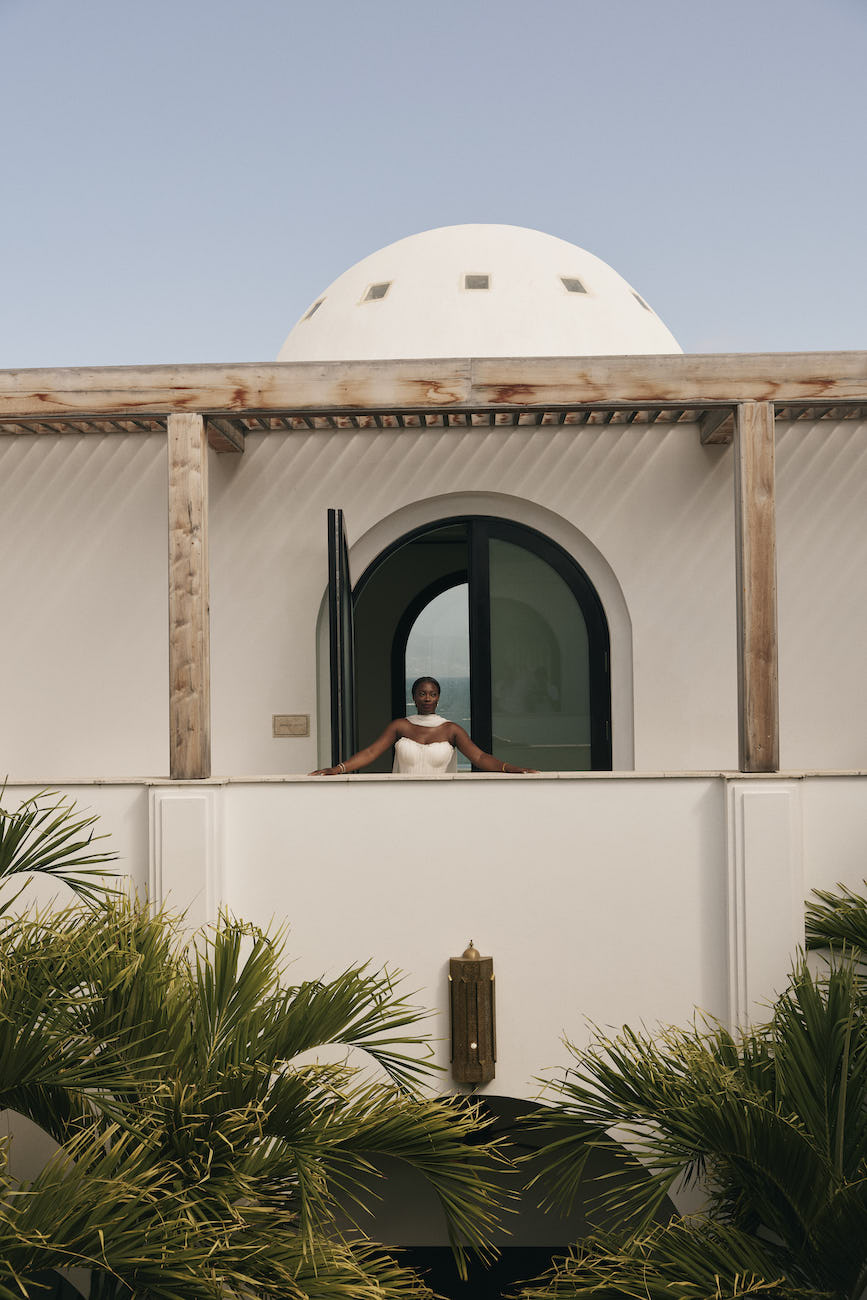 A person in a white dress stands on a balcony in front of an arched doorway with a white dome roof overhead.