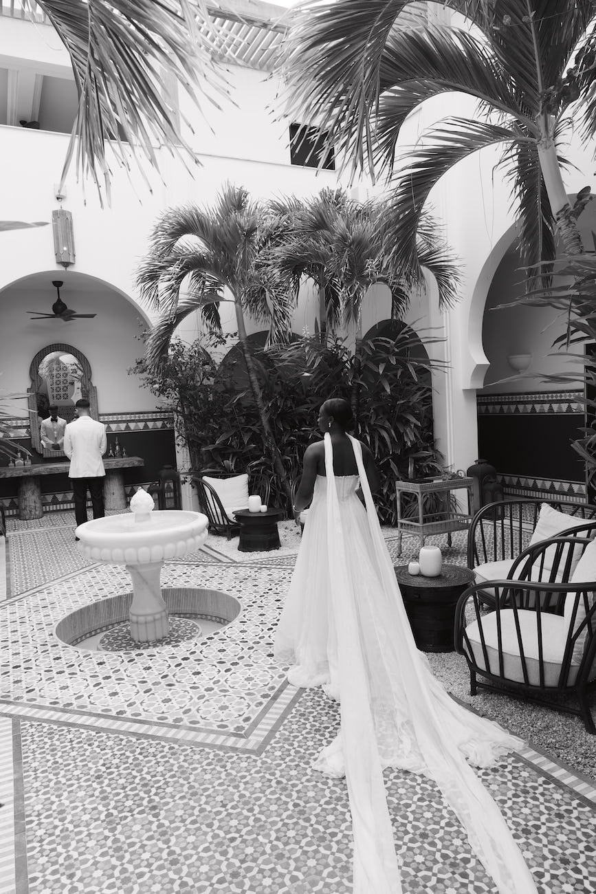 A bride in a long-trained wedding dress walking through a tiled courtyard toward a groom standing by a fountain.