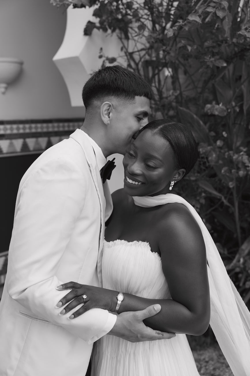 A bride in a strapless gown and a groom in a tuxedo embracing, with the groom kissing the bride on the forehead.