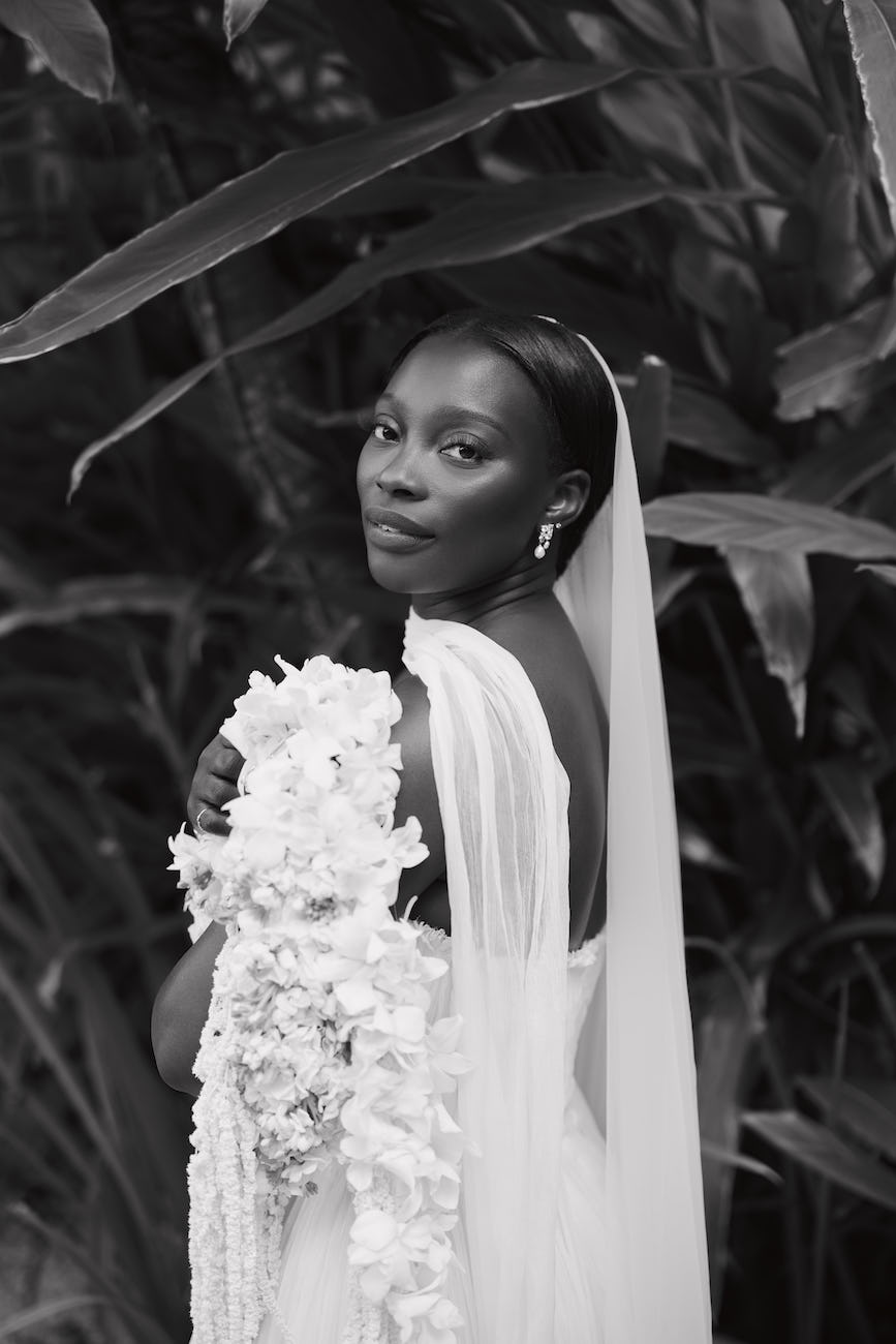 A bride in a veil holds a large white floral bouquet amidst lush foliage.