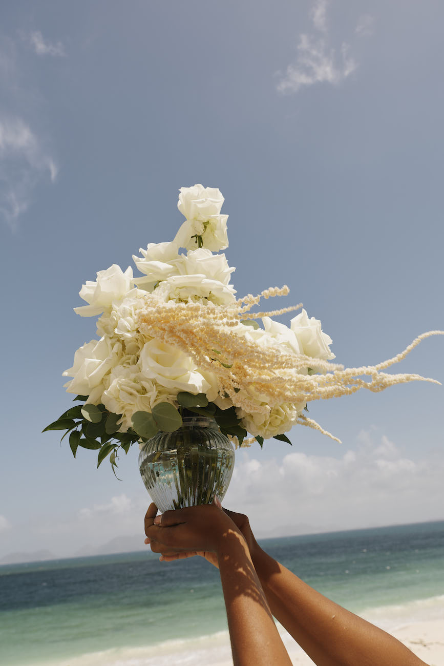 Hands holding a glass vase of white flowers and tall grasses against a beach and ocean background.