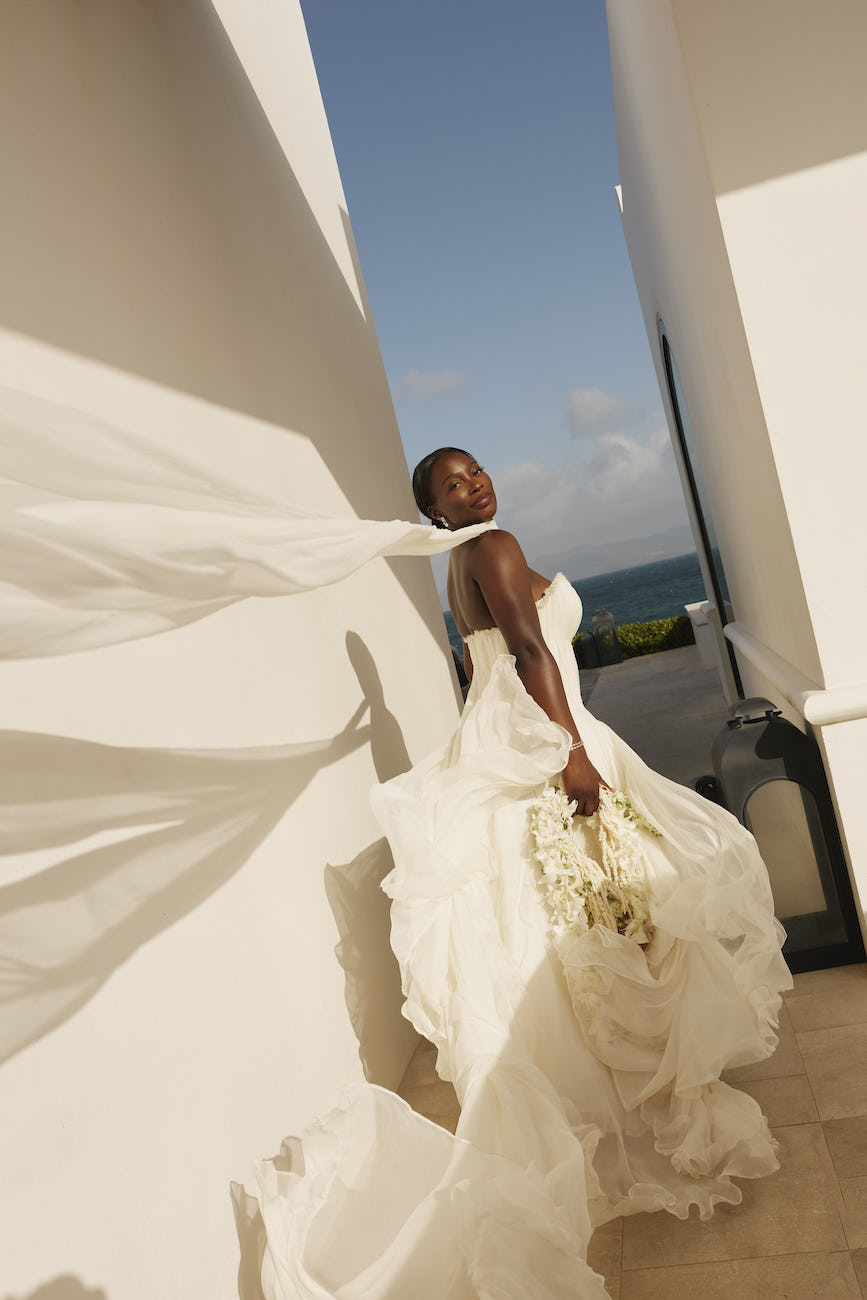 A bride in a white wedding dress holds a bouquet while her veil blows in the wind between two white walls overlooking the ocean.