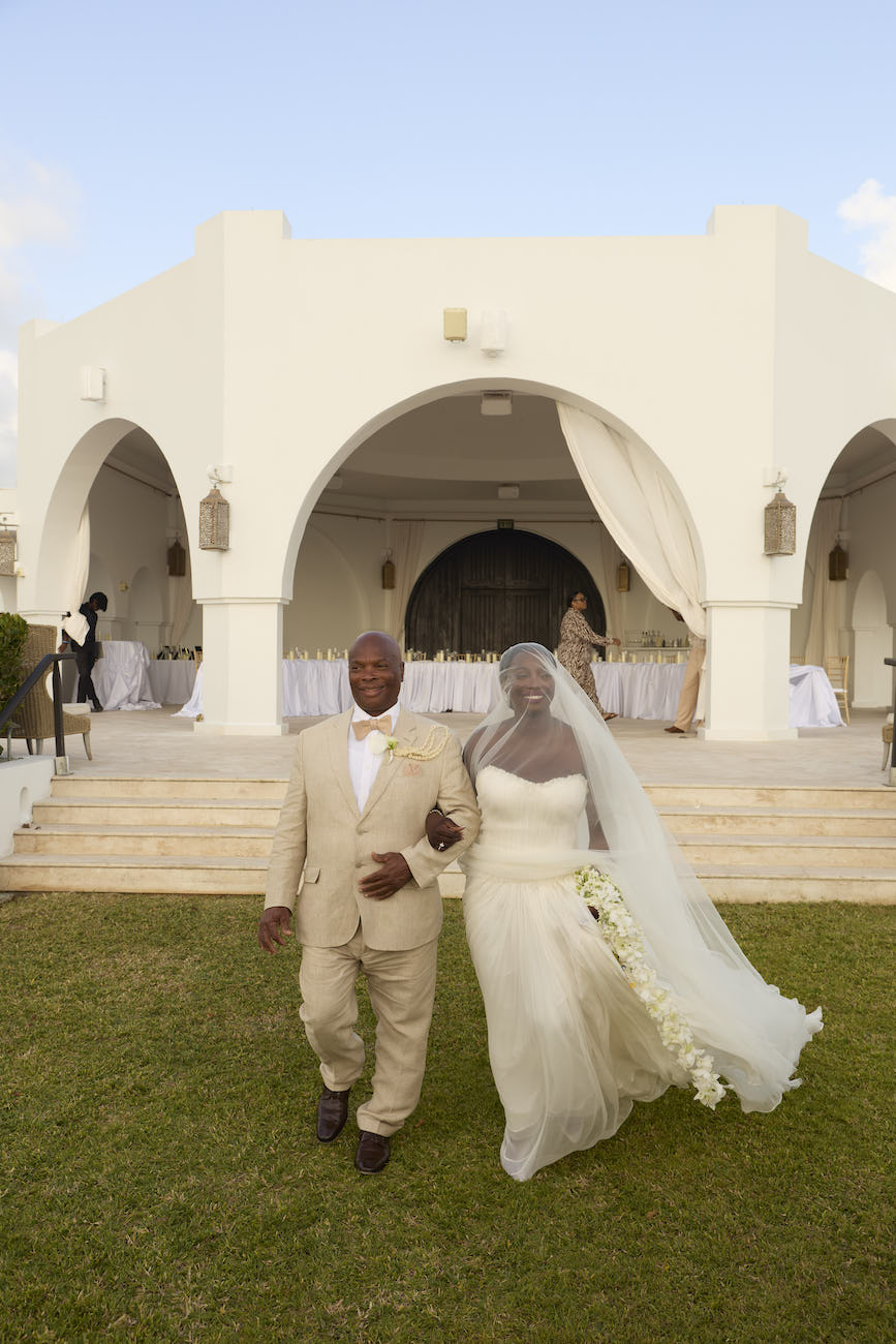 A bride and groom walk hand-in-hand across a lawn in front of an arched building.
