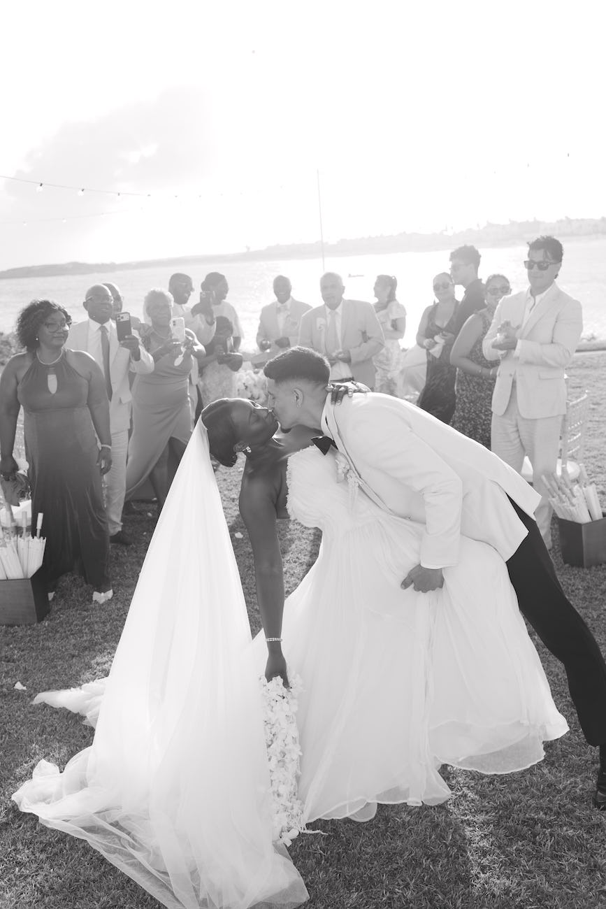 A bride and groom kissing outdoors at a wedding, with guests standing in the background.