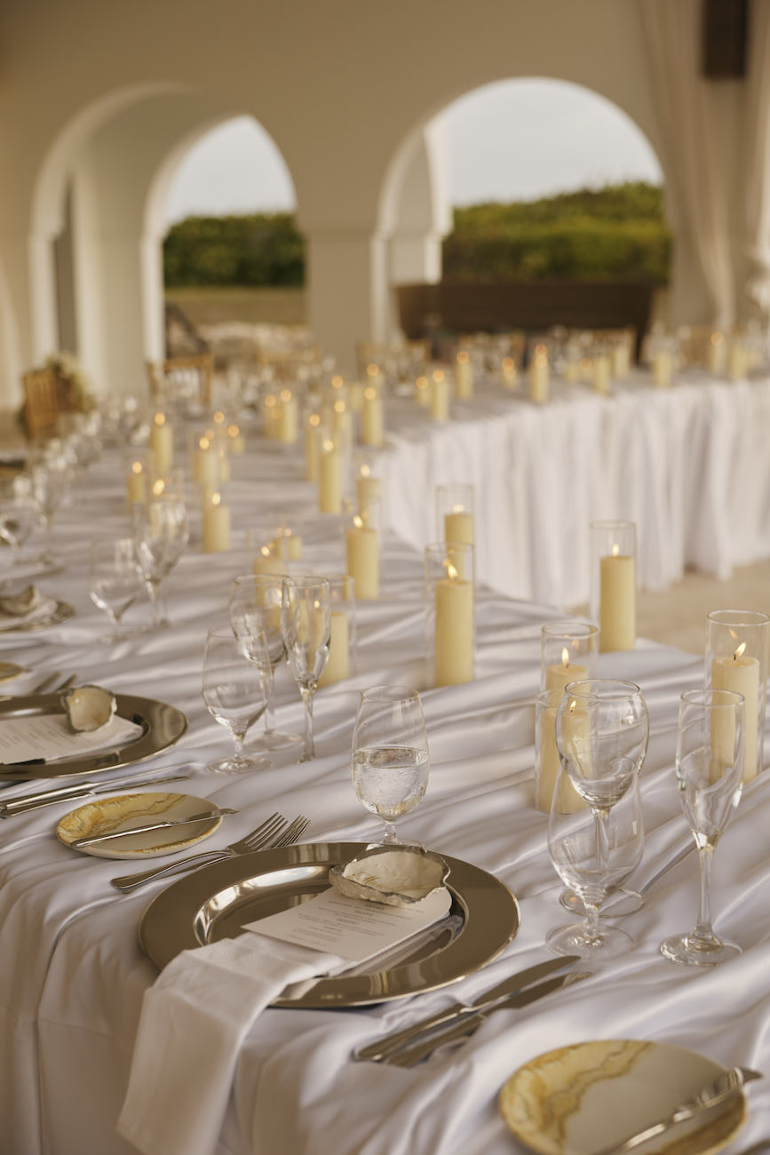 A long banquet table set with white tablecloths, silver plates, wine glasses, and pillar candles in glass holders.
