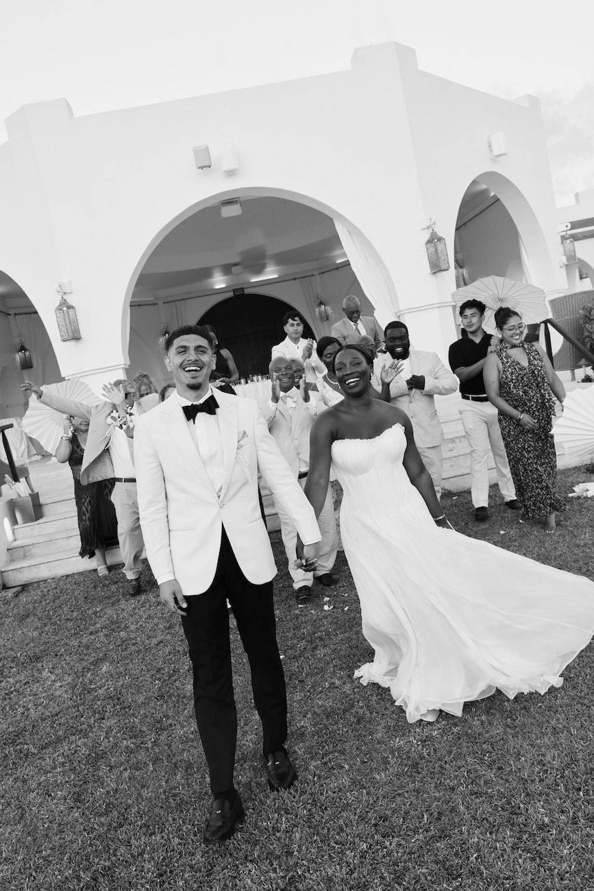 A bride and groom in wedding attire walk hand-in-hand across a lawn with wedding guests in the background.