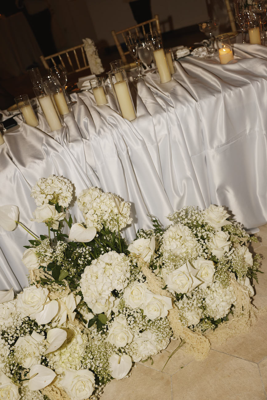 A wedding sweetheart table draped in white fabric, decorated with tall candles and large white floral arrangements on the floor in front.