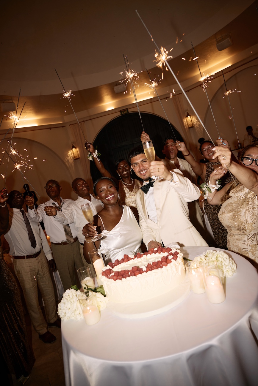 A smiling bride and groom toast with champagne flutes over a round wedding cake topped with raspberries and lit sparklers.