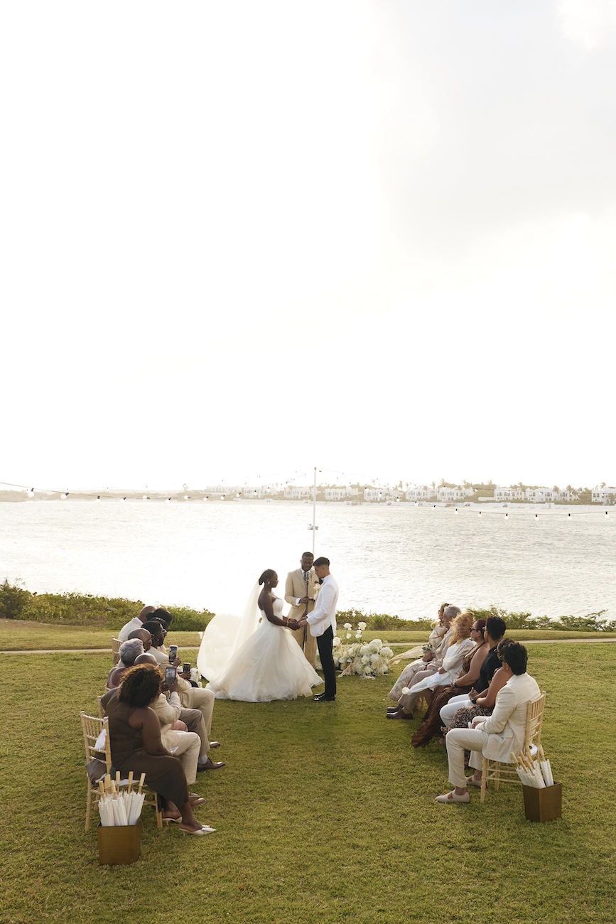 A bride and groom exchange vows at an outdoor wedding ceremony on a lawn overlooking a body of water, with guests seated on either side.