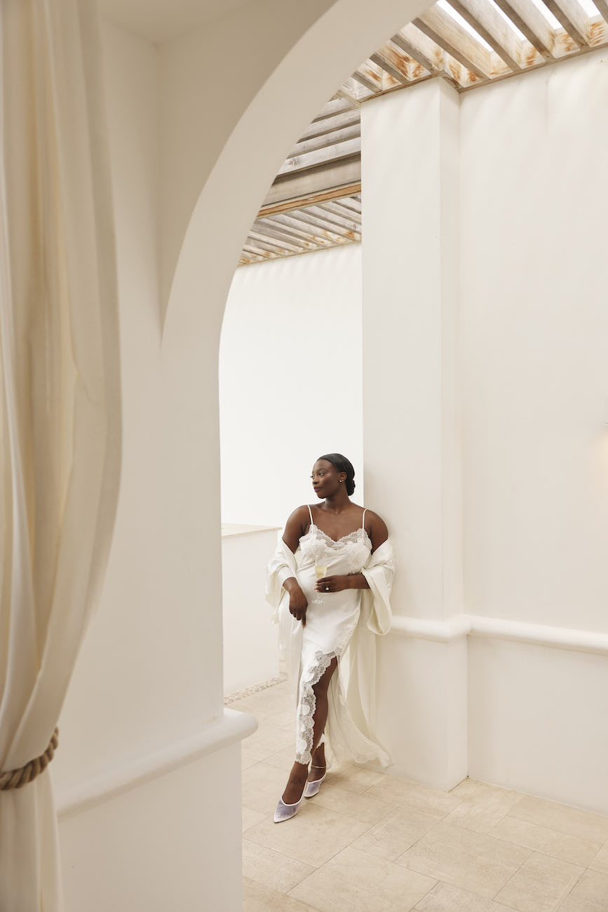 A woman wearing a white silk slip dress and robe stands in an arched stone hallway.