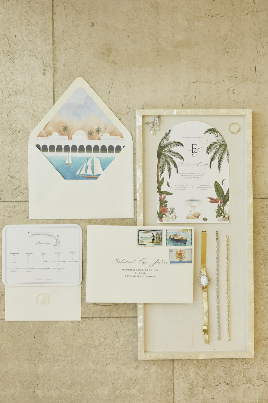 Wedding invitation suite featuring tropical illustrations, an addressed envelope, and a gold wristwatch inside a frame.