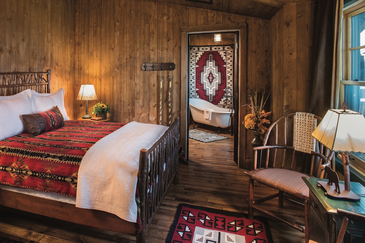 A bedroom with wooden walls featuring a bed with a patterned blanket and a doorway leading to a bathroom with a clawfoot tub.