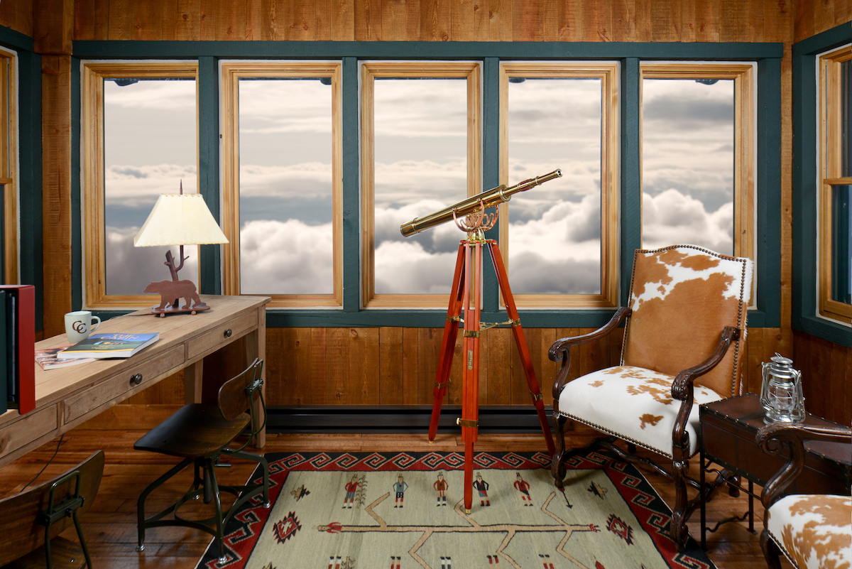 A study with a wooden desk, a brass telescope on a tripod, and an armchair, positioned in front of a row of windows looking out over a bank of clouds.