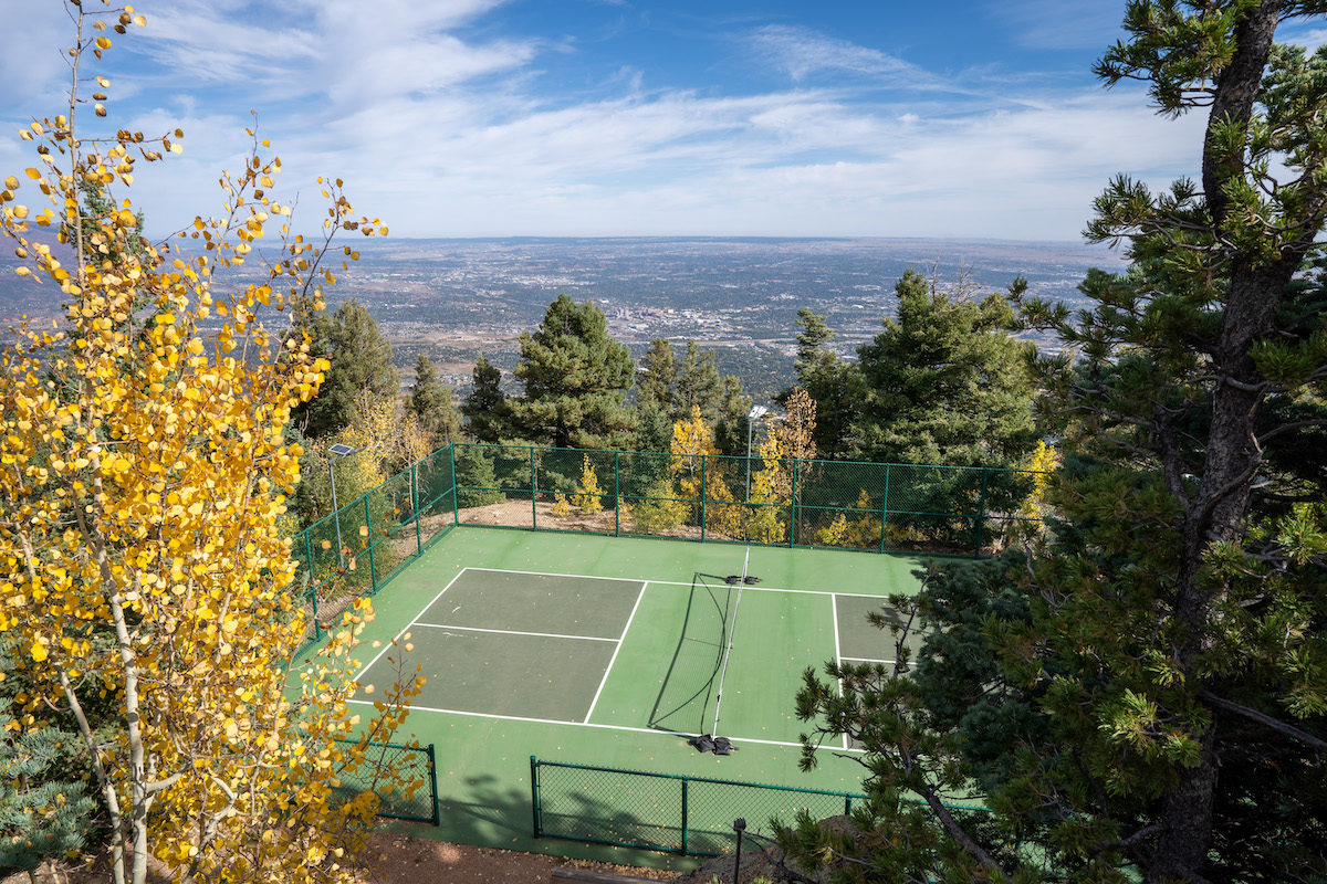 A tennis court surrounded by a fence, viewed from above with a mountain landscape in the distance.