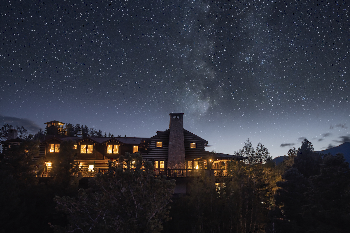 A multi-story lodge lit from within at night, set against a starry sky featuring the Milky Way.