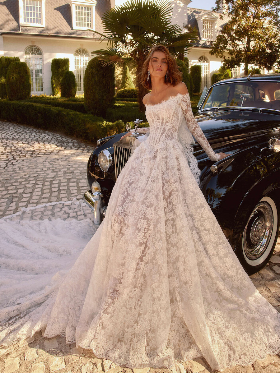 A person in an off-the-shoulder lace wedding gown with a long train standing next to a vintage black car.