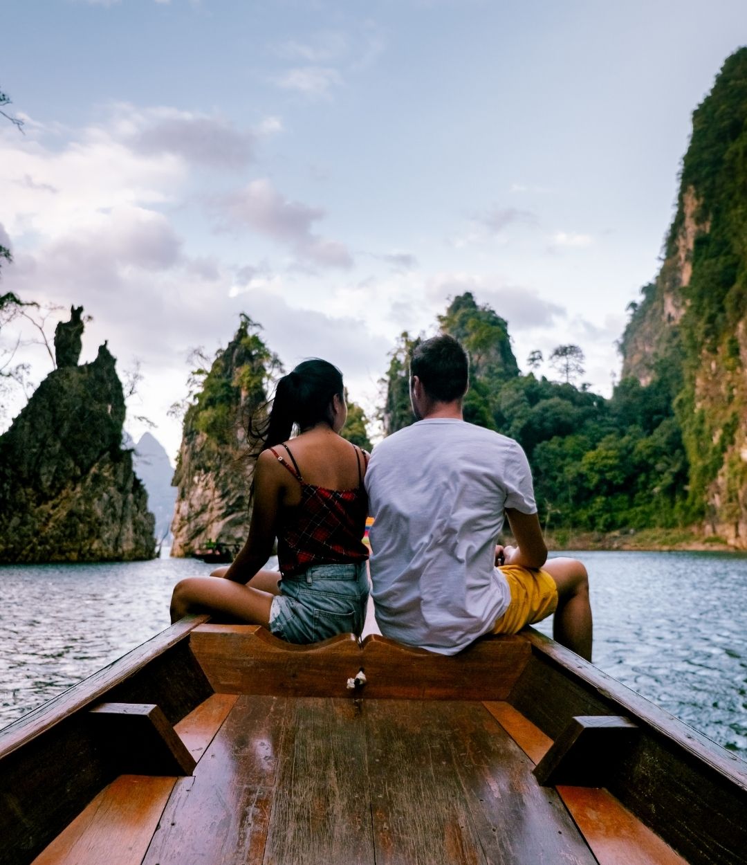 A couple sitting on the front of a wooden longtail boat facing limestone karsts in a lake.