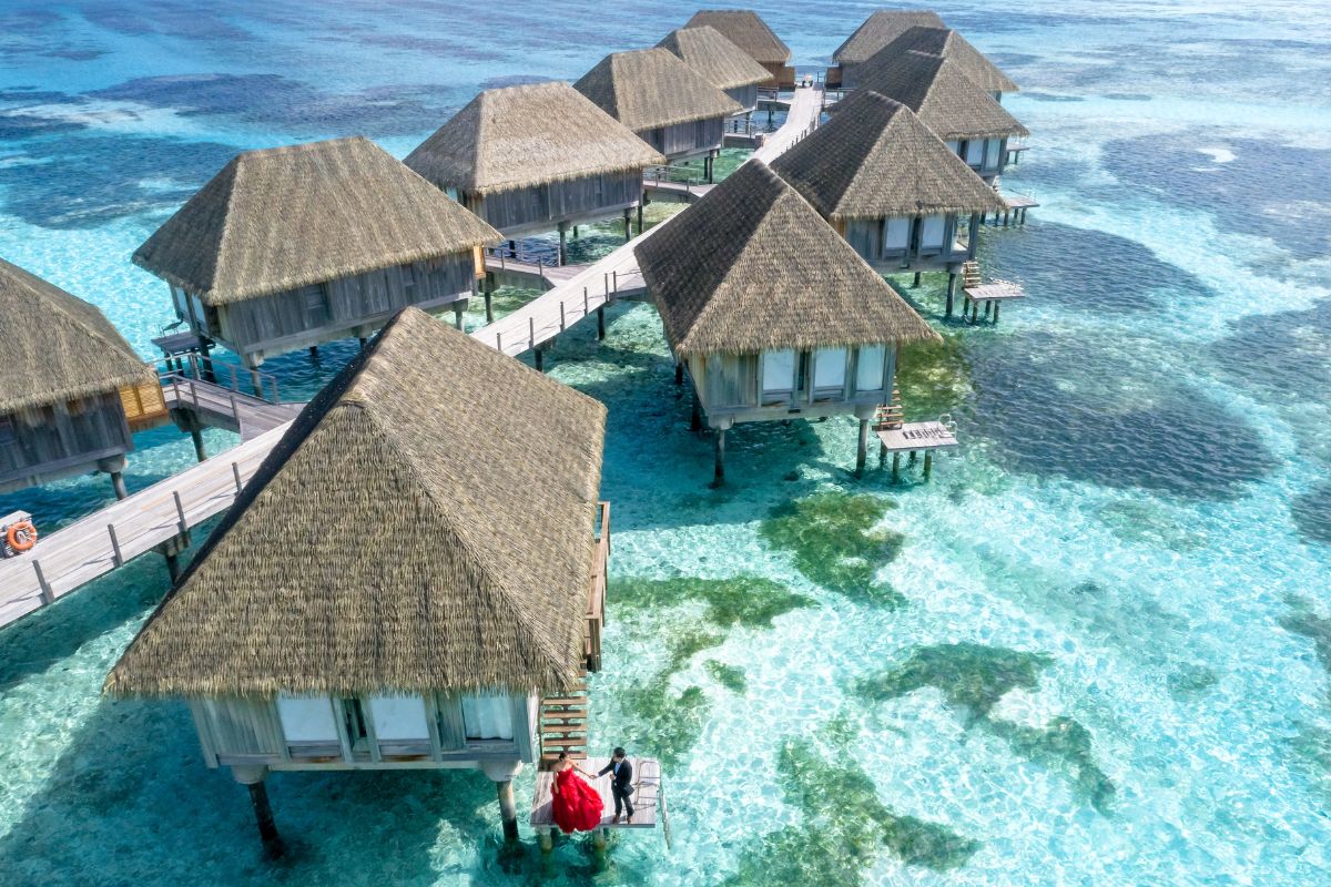 Aerial view of overwater bungalows with thatched roofs connected by wooden walkways over clear turquoise water.