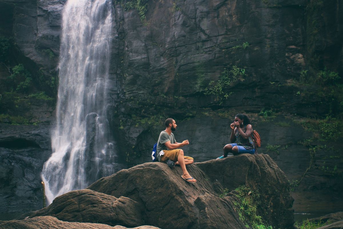 Two people sit on a rock in front of a waterfall.