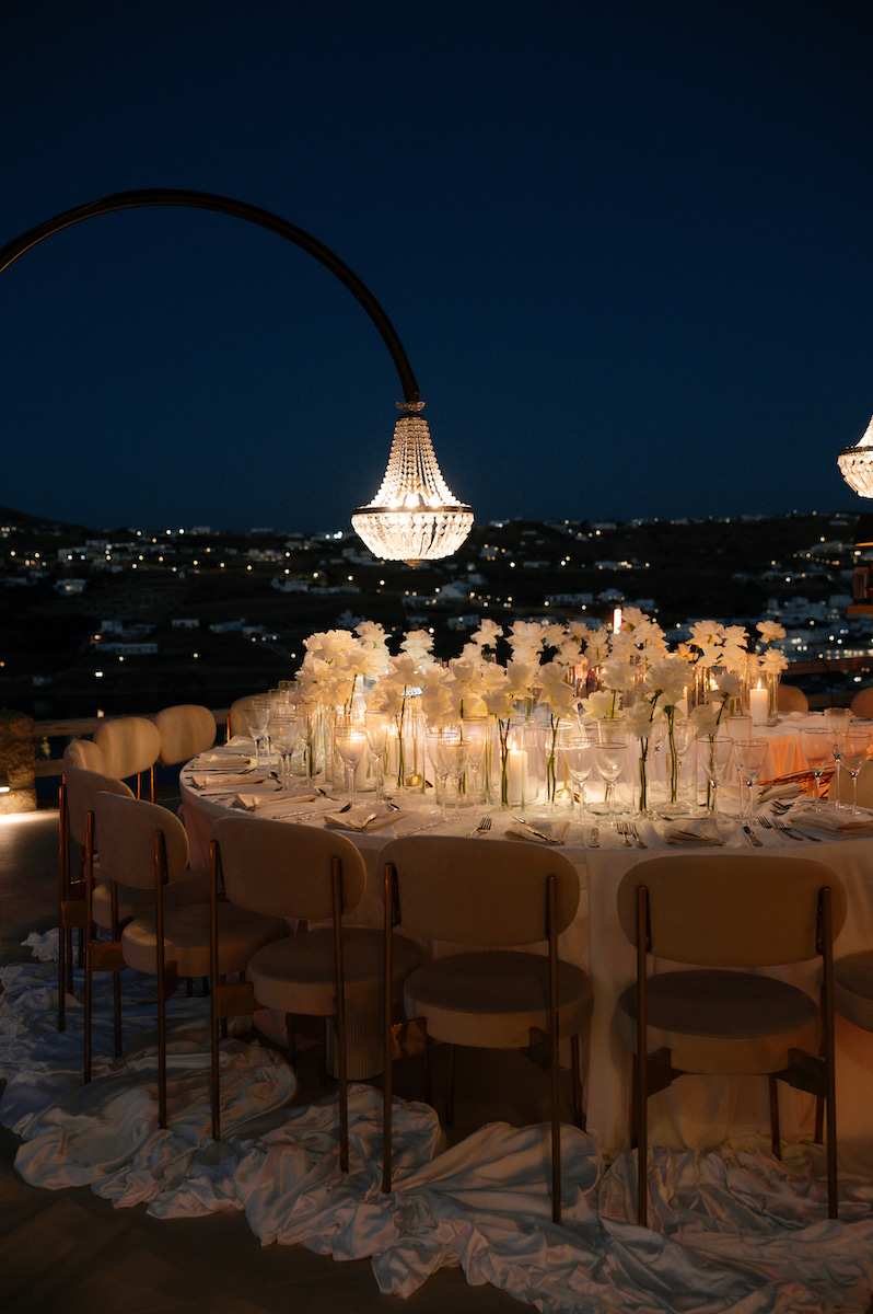 A curved-back dining table set for an evening event, decorated with white floral centerpieces and illuminated by hanging crystal chandeliers against a night sky.
