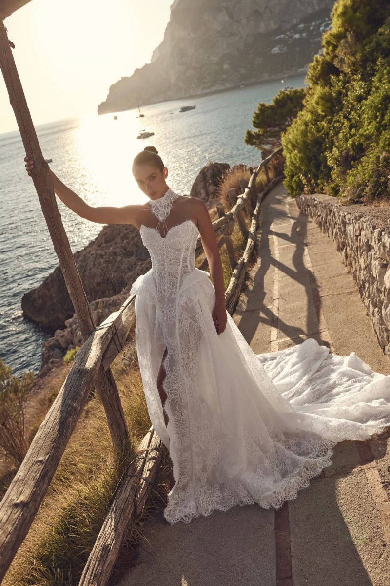 A person wearing a white lace wedding dress with a high collar and a long train standing on a coastal path overlooking the sea.