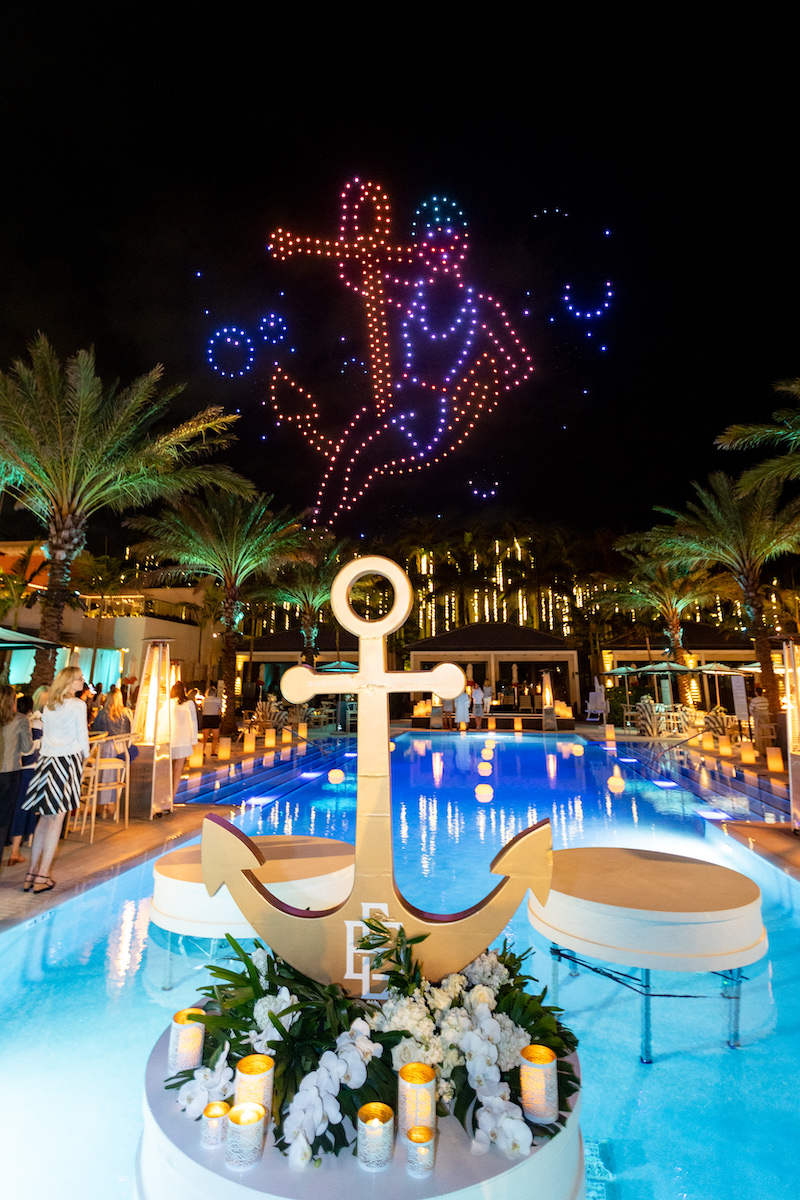 A large anchor prop on a pool platform in front of a drone light show featuring an anchor and a sailor figure above a swimming pool at night.