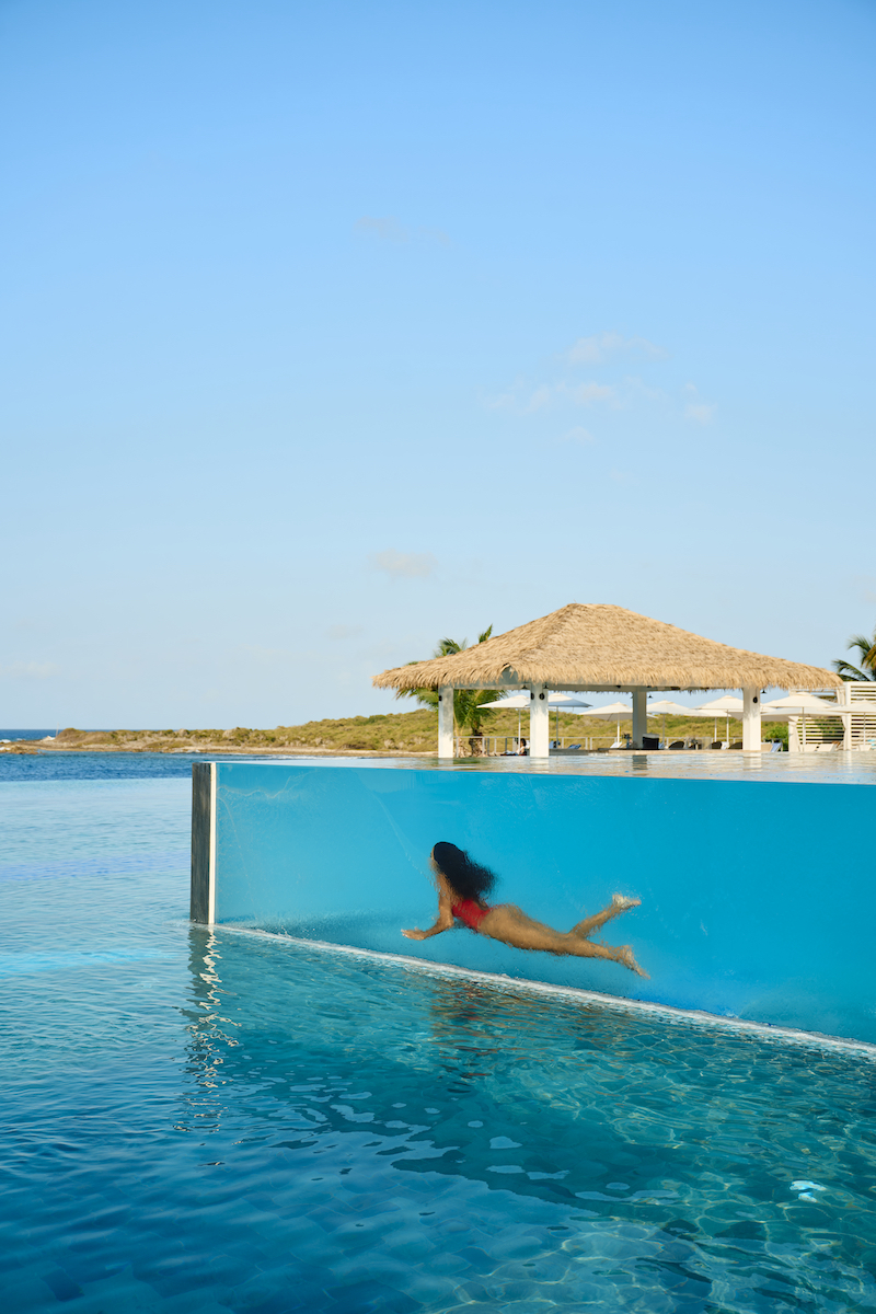 A person in a red swimsuit swims underwater in a glass-walled infinity pool.