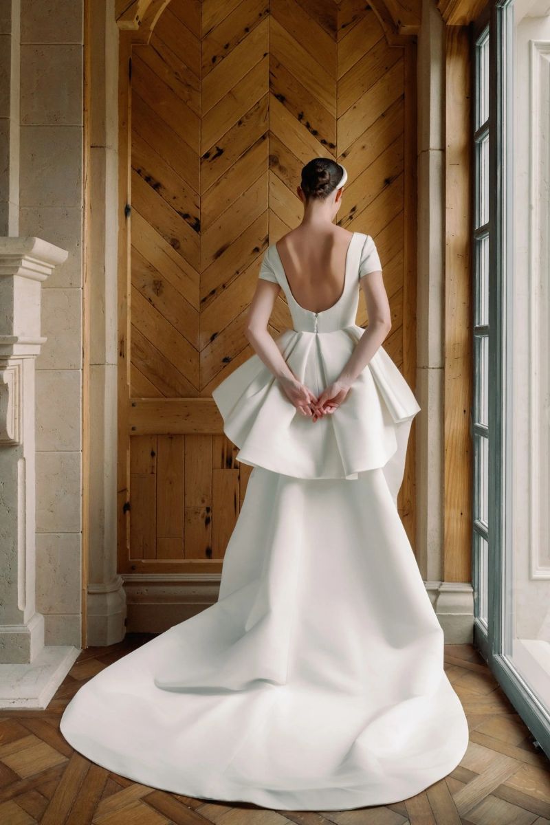 A person seen from behind wearing a white wedding gown with a scoop back and a flared peplum skirt with a train.