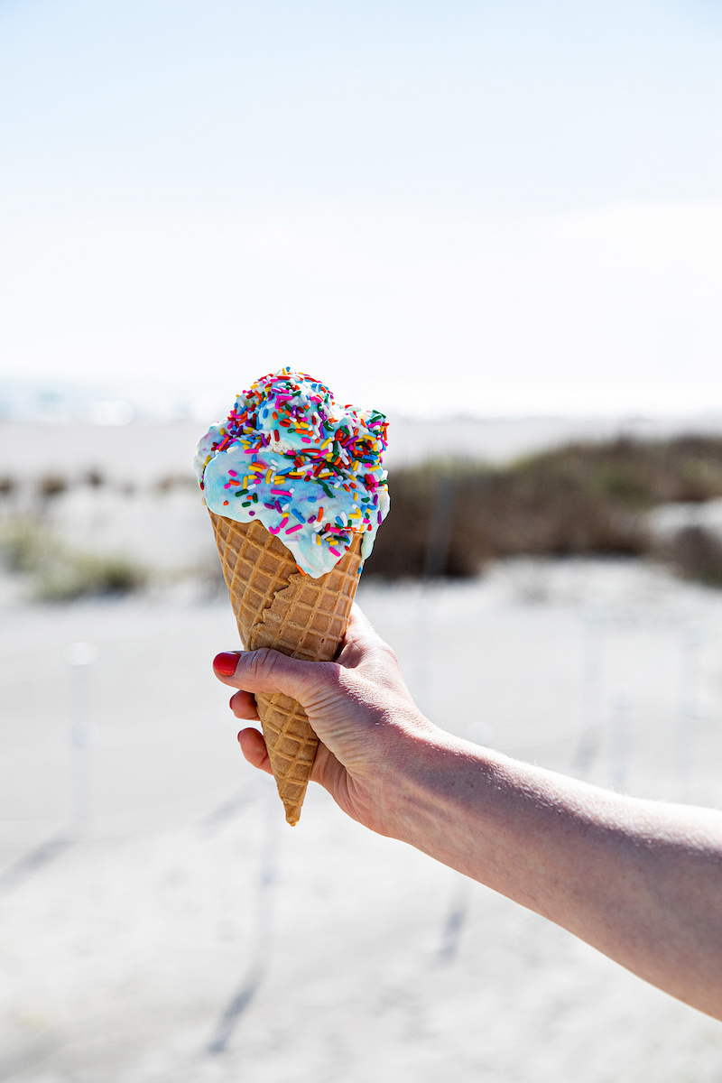 A hand holds a waffle cone topped with blue ice cream covered in rainbow sprinkles at a beach.