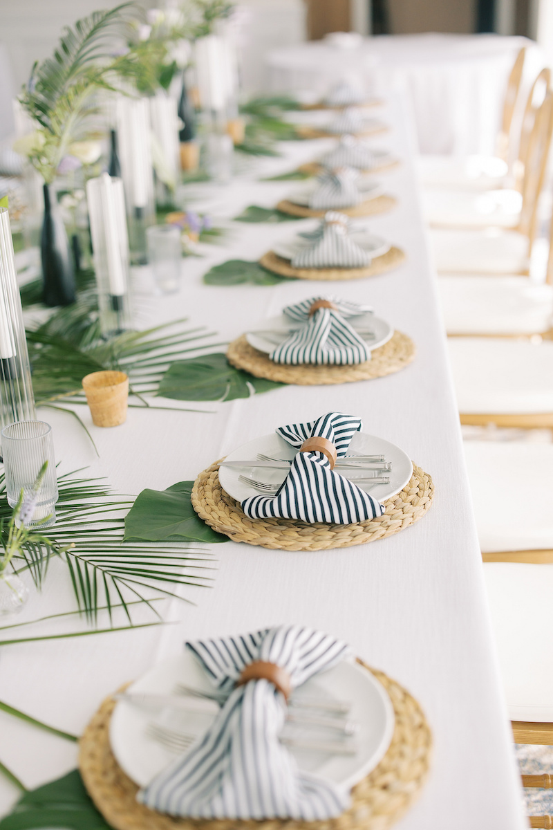Long banquet table set with white plates on woven placemats, topped with blue and white striped napkins held by wooden rings and decorative palm leaves.
