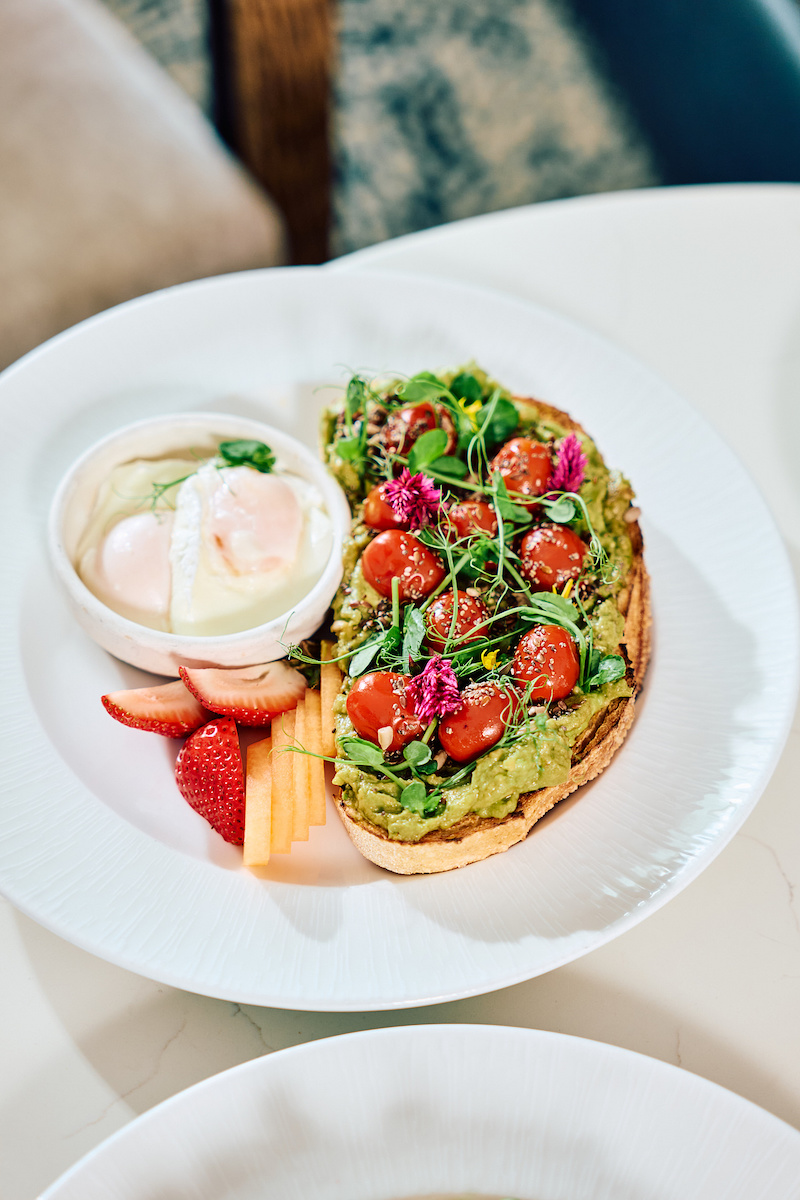 Avocado toast topped with cherry tomatoes and microgreens, served with a side of poached eggs, sliced strawberries, and carrot sticks.