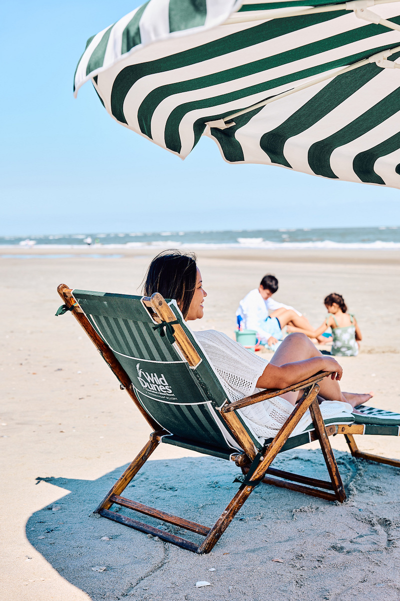 A person sits in a green striped beach chair under a matching umbrella, looking toward two people playing in the sand in the distance.