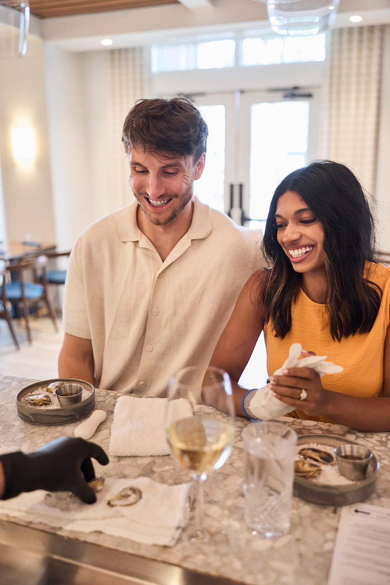 A smiling man and woman sit at a bar counter while someone in black gloves places an oyster on a napkin in front of them.